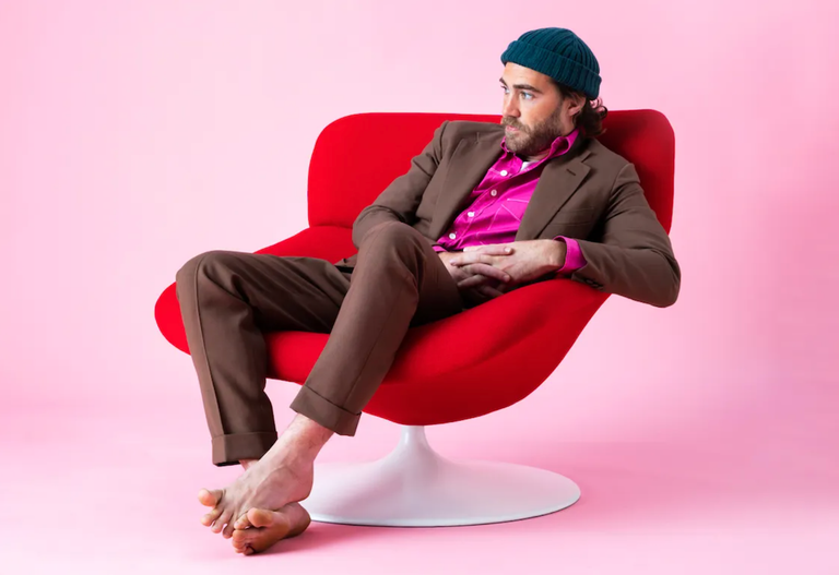 Australian musician Matt Corby sitting barefoot in a red chair wearing a brown suit and beanie for a studio promo artist portrait photo shoot.